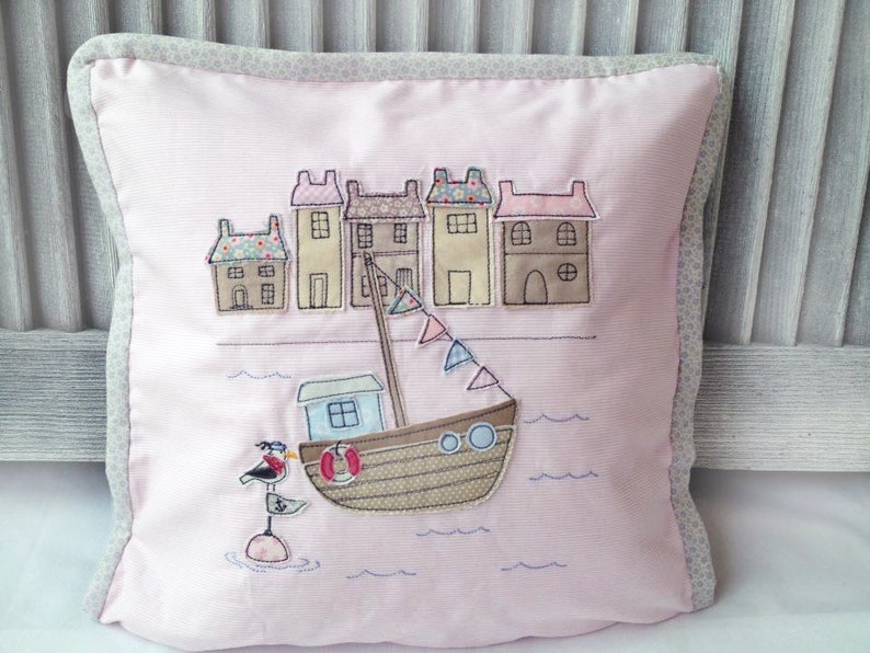May include: A pink pillow with a white border features an embroidered design of a small town with houses and a boat on the water. The boat has a life preserver and a seagull perched on a buoy.