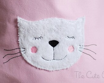May include: A pink fabric bag with a white, fluffy, embroidered cat face. The cat has closed eyes, a black nose, and pink cheeks.