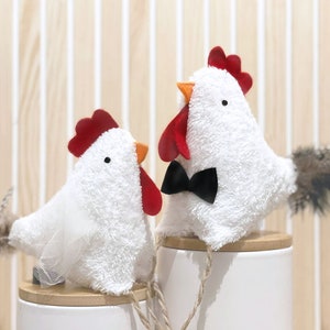May include: Two white plush chickens, one wearing a white veil and the other a black bow tie, are sitting on top of white containers with wooden lids. The chickens have red combs and beaks.