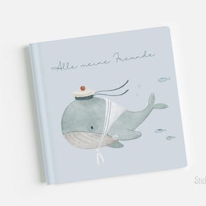 Op de afbeelding: Een lichtblauw boek met de tekst "Alle meine Freunde" op de omslag. De omslag toont een cartoonillustratie van een blauwe walvis die een matrozenhoed en een witte matrozenkraag draagt, zwemmend met kleine vissen.