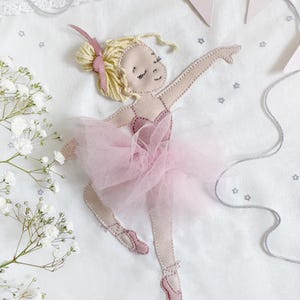 May include: A pink and white embroidered ballerina applique with a pink tulle skirt and a pink bow in her hair. The ballerina is on a white background with silver stars.