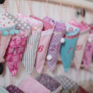 May include: A collection of colorful fabric cones, each with a unique pattern and number, hanging from a white wall. The cones are in shades of pink, purple, and blue, with polka dots, stripes, and floral designs. Some have pom-poms or lace trim.