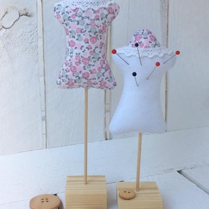 May include: Two fabric pincushions on wooden stands. The pincushion on the left is pink with a floral pattern and a white lace trim. The pincushion on the right is white with a white lace trim and has red and black pins sticking out of it. Both pincushions are on square wooden bases with a button on each base.