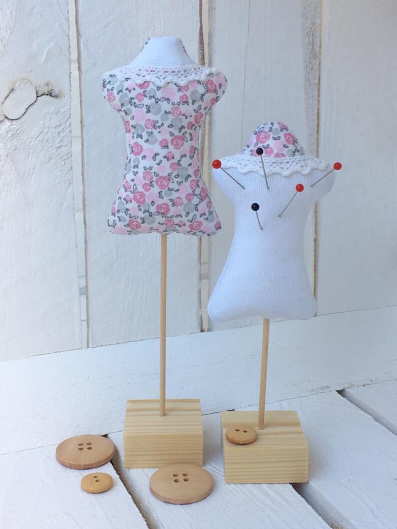 May include: Two fabric pincushions on wooden stands. The pincushion on the left is pink with a floral pattern and a white lace trim. The pincushion on the right is white with a white lace trim and has red and black pins sticking out of it. Both pincushions are on square wooden bases with a button on each base.