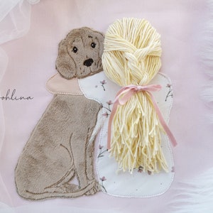 May include: A pink fabric applique featuring a brown dog and a blonde braided doll with a pink bow. The text "Johllina" is embroidered on the fabric.