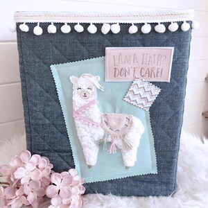 May include: A blue quilted storage bin with a white pom-pom trim. The bin features a white fluffy llama applique with pink accents and a pink patch that says "Llama Hair? Don't Care!"