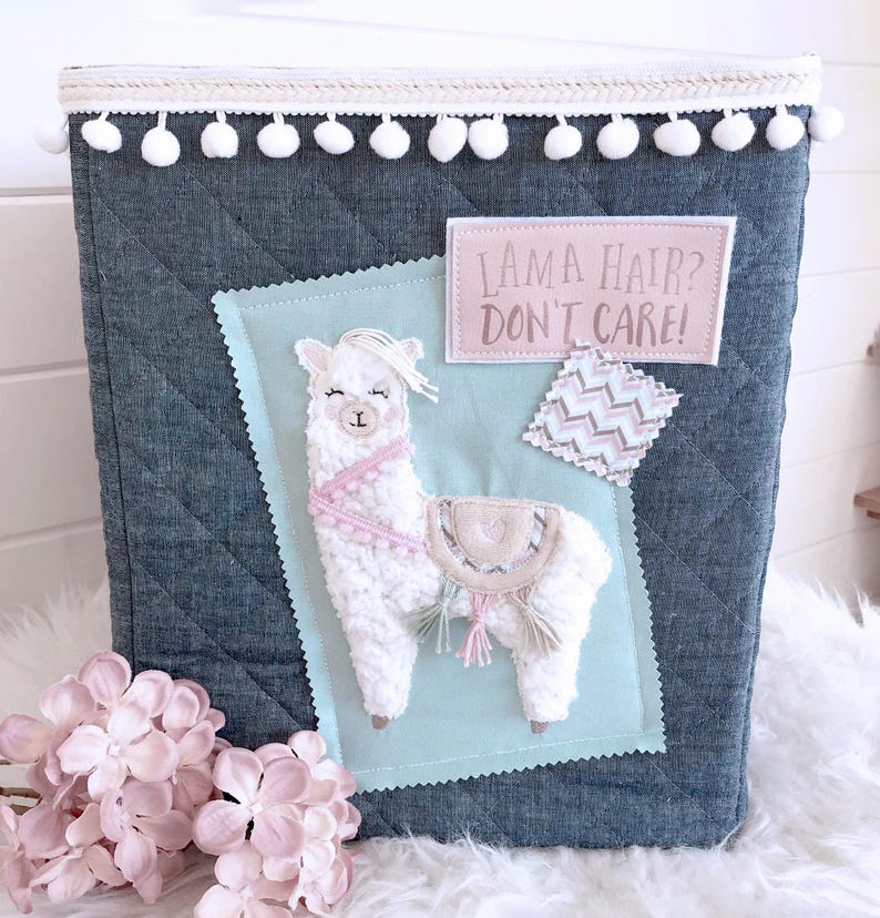 May include: A blue quilted storage bin with a white pom-pom trim. The bin features a white fluffy llama applique with pink accents and a pink patch that says "Llama Hair? Don't Care!"