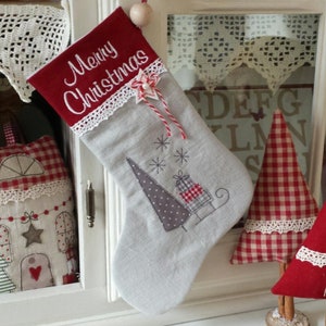 Puede incluir: Una media de Navidad gris y roja con ribete de encaje blanco y las palabras "Merry Christmas" bordadas en el puño rojo. La media está decorada con un árbol gris y blanco y un trineo con regalos.