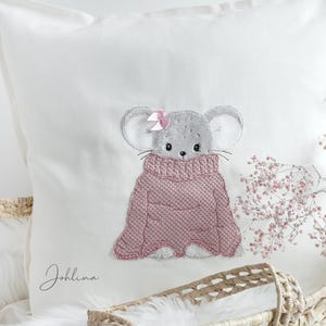 Könnte beinhalten: Ein weißes Kissen mit einer rosa und grauen gestickten Maus, die einen rosa Pullover und eine rosa Schleife trägt. Das Kissen trägt den Namen "Johllina" gestickt.