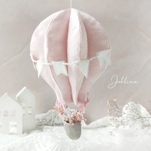 May include: A pink fabric hot air balloon with white flags hanging from the bottom. The balloon is suspended from the ceiling and has a small basket with white flowers.