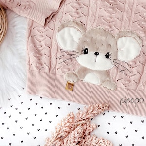 Puede incluir: Un suéter de punto rosa con una aplicación de ratón marrón. El suéter tiene un patrón de punto de cable y el ratón tiene una cara blanca con ojos negros y una nariz negra. El suéter tiene la palabra "pippa" bordada en la parte inferior.