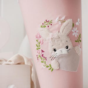 Könnte beinhalten: Hellrosa Stoffkegel mit einem gestickten Hasen-Design. Der Hase hält ein graues Herz und ist von einem Blumenkranz mit rosa und weißen Blüten umgeben. Der Hase hat eine Schleife auf dem Kopf.