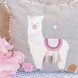May include: A gray pillow with a white llama applique. The llama has pink details and a pink heart on its saddle. The pillow has a pink pom-pom trim and a hand-made tag.
