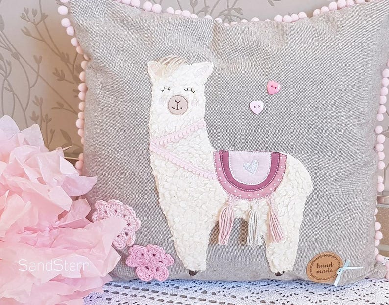 May include: A gray pillow with a white llama applique. The llama has pink details and a pink heart on its saddle. The pillow has a pink pom-pom trim and a hand-made tag.