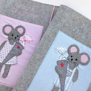 Könnte beinhalten: Zwei graue Filzordner mit gestickten Mäusen, die einen kegelförmigen Leckerbissen halten. Die Ordner haben rosa und blaue Gingham-Stofftaschen mit dem Text "Meine Zeugnisse" auf der Vorderseite bestickt.
