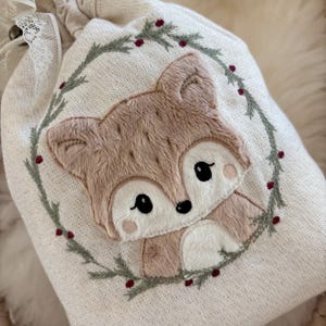 Puede incluir: Una bolsa beige con cordón y un diseño bordado de zorro detallado. El zorro está rodeado por una corona verde con bayas rojas. La bolsa tiene un cierre de encaje y botón, ideal para regalos.