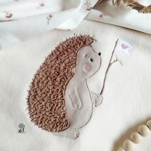 Op de afbeelding: Een wit pluche deken met een geborduurde bruine egel die een witte vlag met een roze hart vasthoudt. Het deken heeft een klein bloemenpatroon.