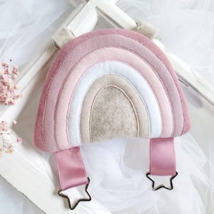 May include: A pink and beige rainbow-shaped plush toy with two pink ribbon loops and star-shaped metal clips.