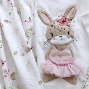 May include: A white fabric with a floral pattern featuring a brown embroidered bunny wearing a pink tutu and a flower crown.