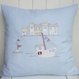 May include: A blue and white striped pillow with an embroidered design of a boat with a red roof and a lighthouse with a bird on top. The boat is sailing past a row of houses with red roofs.
