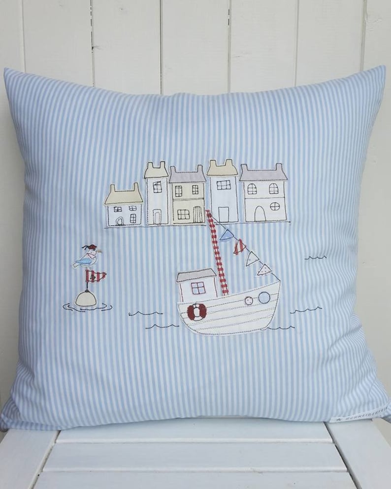 May include: A blue and white striped pillow with an embroidered design of a boat with a red roof and a lighthouse with a bird on top. The boat is sailing past a row of houses with red roofs.