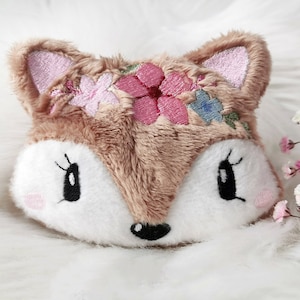 Può includere: Un giocattolo di peluche a forma di volpe con fiori ricamati sulla testa. La volpe ha un viso marrone e bianco con occhi neri e un naso nero.