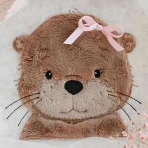 May include: A brown plush otter with a pink bow on its head. The otter has embroidered eyes, a nose, and whiskers.