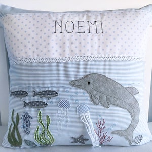 May include: A blue and white pillow with a dolphin, fish, jellyfish, and seaweed embroidered on it. The pillow has a white lace trim and the name "NOEMI" embroidered on the top.