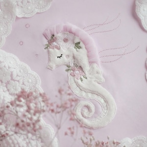 May include: A pink fabric with a white lace border and a white embroidered seahorse with pink flowers. The seahorse has a pink tulle mane and tail.