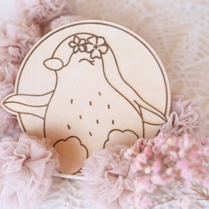 May include: A wooden penguin with a flower crown, painted in a light brown color. The penguin is standing on a white background with pink tulle and flowers.