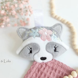 Könnte beinhalten: Ein grauer und weißer Plüschwaschbären mit gestickten rosa und blauen Blumen auf dem Kopf. Der Waschbär ist an einem rosa Waffelgewebe befestigt. Der Text "Mili & Lola" ist in der linken unteren Ecke des Bildes sichtbar.