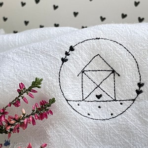 Pode incluir: Tecido branco com um desenho bordado preto de uma casa dentro de um círculo. A casa tem um coração na parte inferior e o círculo é rodeado por pequenos corações.