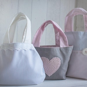 Op de afbeelding: Drie kleine draagtassen met verschillende patronen. De eerste tas is lichtblauw met witte strepen en witte kantversiering. De tweede tas is grijs met witte stippen en een roze hart. De derde tas is roze en wit geruit met een witte knoop.