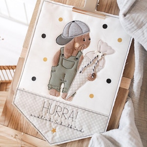 Pode incluir: Uma faixa decorativa com um coelho de desenho animado usando macacão e boné, segurando um presente em forma de cenoura. A faixa tem o texto "HURRA! endlich Schulkind" e é decorada com bolinhas pretas e douradas.