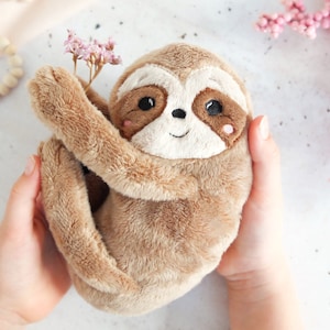 Puede incluir: Un peluche de perezoso en tonos marrones y beige, con rasgos faciales bordados y una pequeña flor rosa. El perezoso es sostenido por manos. Tiene un aspecto suave y texturizado.