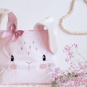 Peut inclure: Un panier de rangement en forme de lapin à rayures roses et blanches avec un nœud rose et des oreilles en fourrure blanche.