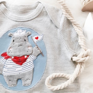 May include: A grey and white striped baby onesie with a blue and white appliqué of a hippo wearing a sailor hat and holding a red heart flag.