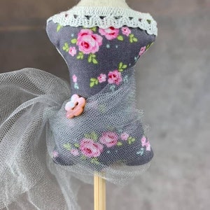 May include: A gray fabric mannequin with a floral print and a white lace collar. The mannequin is wearing a sheer gray tulle skirt and has a pink button on the bodice. The mannequin is on a wooden stick.