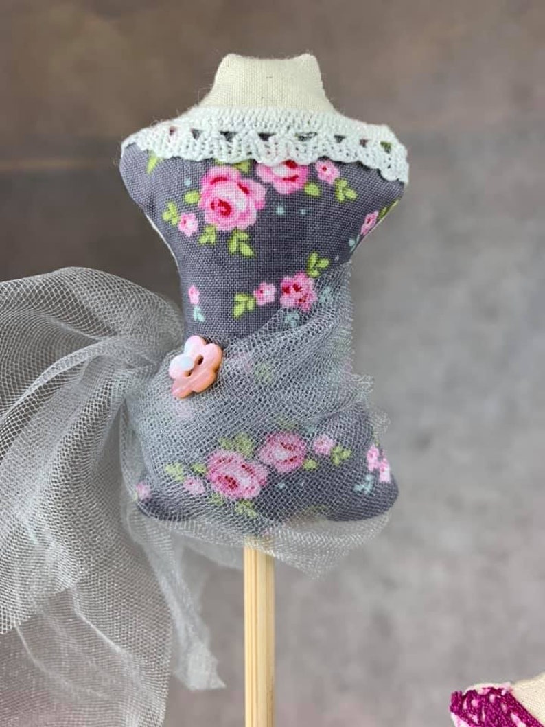 May include: A gray fabric mannequin with a floral print and a white lace collar. The mannequin is wearing a sheer gray tulle skirt and has a pink button on the bodice. The mannequin is on a wooden stick.