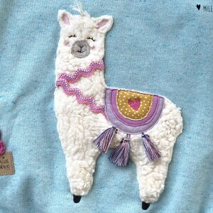 May include: A light blue sweatshirt with a white fluffy llama applique. The llama has a pink and white saddle with a small heart on it. The llama has a pink and white beaded necklace. The sweatshirt has the text "Little Love" on the bottom left corner.