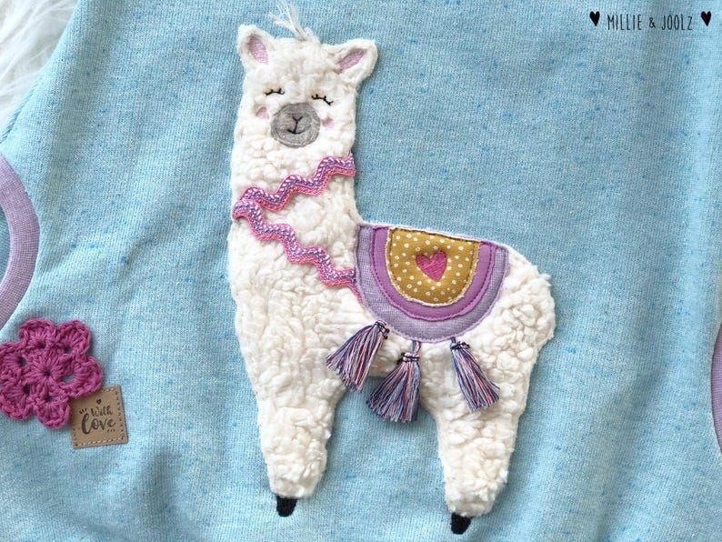 May include: A light blue sweatshirt with a white fluffy llama applique. The llama has a pink and white saddle with a small heart on it. The llama has a pink and white beaded necklace. The sweatshirt has the text "Little Love" on the bottom left corner.