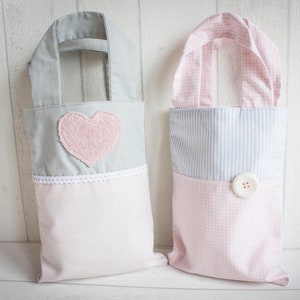 May include: Two fabric tote bags with pink and blue patterns. One bag has a pink heart applique and the other has a white button. Both bags have white handles.