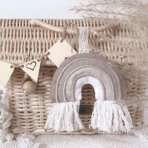 May include: A beige and white rainbow with fringe hanging from the bottom. The rainbow is made of fabric and is hanging from a wicker basket with a wooden heart banner.