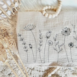 May include: A beige muslin baby blanket with black embroidered wildflowers. The blanket has a subtle textured pattern and is perfect for a nursery or as a gift.