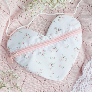Könnte beinhalten: Eine herzförmige Geldbörse mit weißem Blumenmuster und einem rosa Reißverschluss. Die Geldbörse hat einen passenden rosa Riemen und liegt auf einer rosa Strickdecke. Zarte weiße Blumen sind um die Geldbörse gelegt.