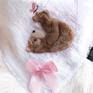 May include: A white baby blanket with a brown bear applique, a pink butterfly applique, and a pink bow. The blanket has a white lace trim and a black tag with the text "Jolina".