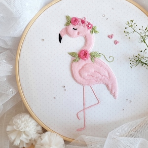 May include: Pink flamingo embroidered on white fabric with pink flowers and green leaves. The embroidery is in a wooden hoop.
