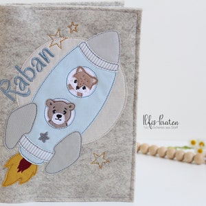 Puede incluir: Una cubierta de libro de fieltro gris con un diseño de cohete. El cohete es azul y blanco con una llama amarilla. La cubierta del libro tiene el nombre "Raban" bordado. La cubierta del libro está decorada con estrellas.