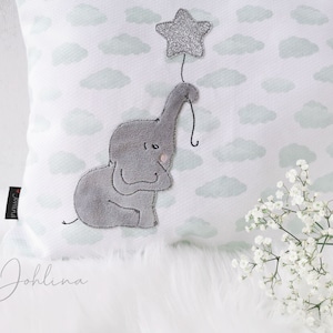 Puede incluir: Un cojín blanco con un estampado de nubes celeste. Un aplique de elefante gris sujeta una estrella plateada con purpurina. El cojín tiene el texto "Johlina" en cursiva.