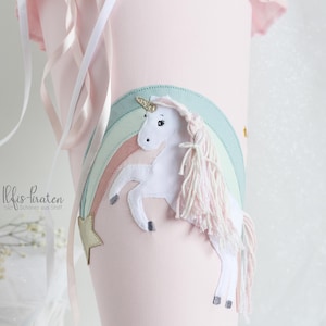 Puede incluir: Un unicornio rosa y blanco con una crin y cola de arcoíris, de pie sobre un arcoíris con una estrella dorada. El unicornio está sobre un fondo rosa con una cinta blanca.